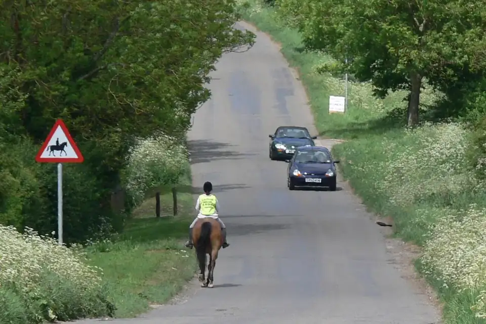 Une cavalière en gilet jaune sur une petite route de campagne bordée de haies, croisée par deux voitures, un panneau de passage de cavaliers sur le bas-côté